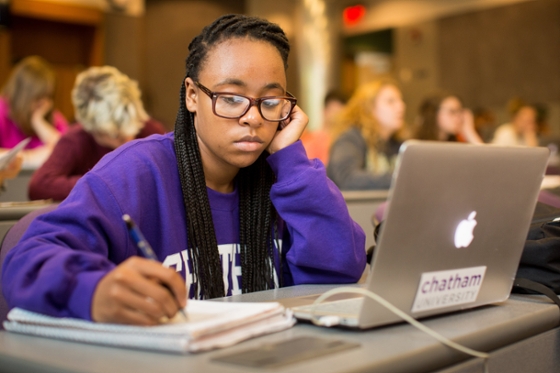 Photo of a student in a Chatham University sweatshirt, working at a laptop in a lecture hall and taking notes