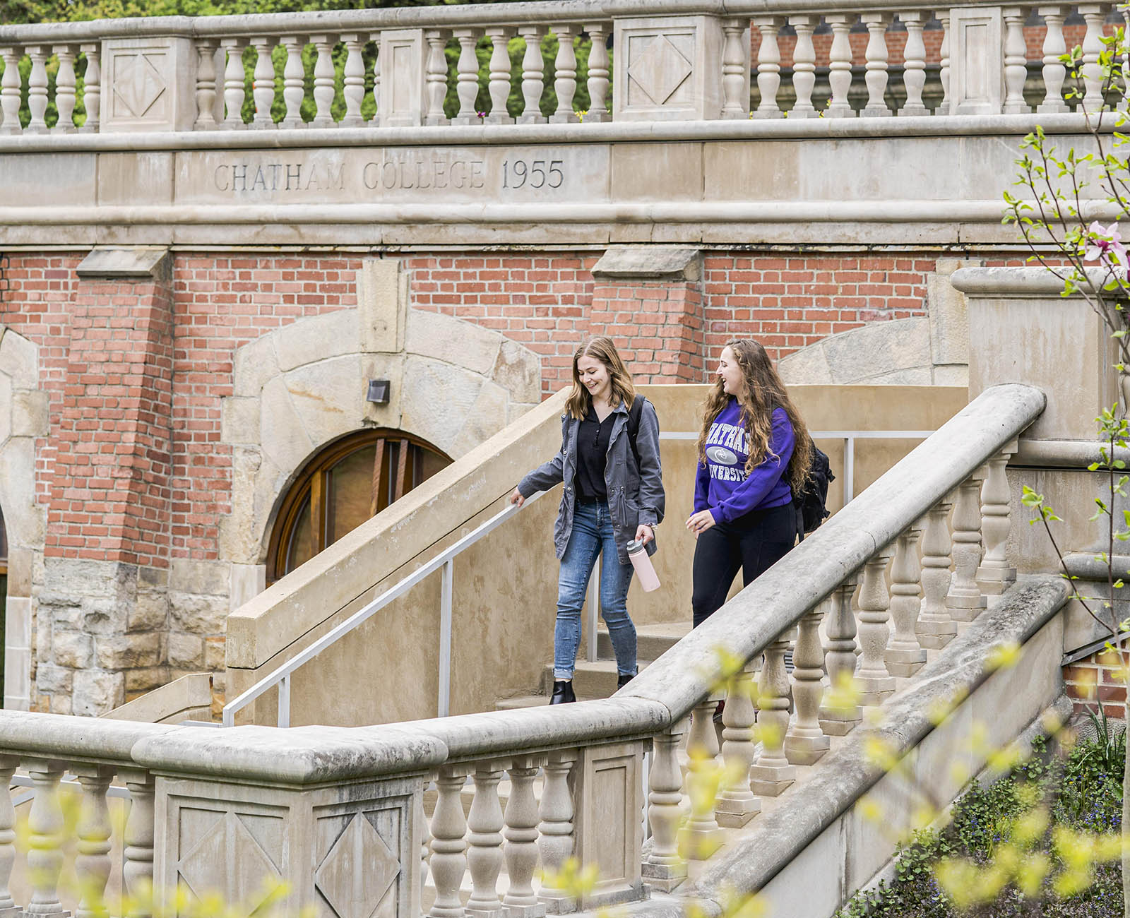 Photo of two female college students walking down the staircase of a redbrick academic building outside on Chatham University's Shadyside Campus
