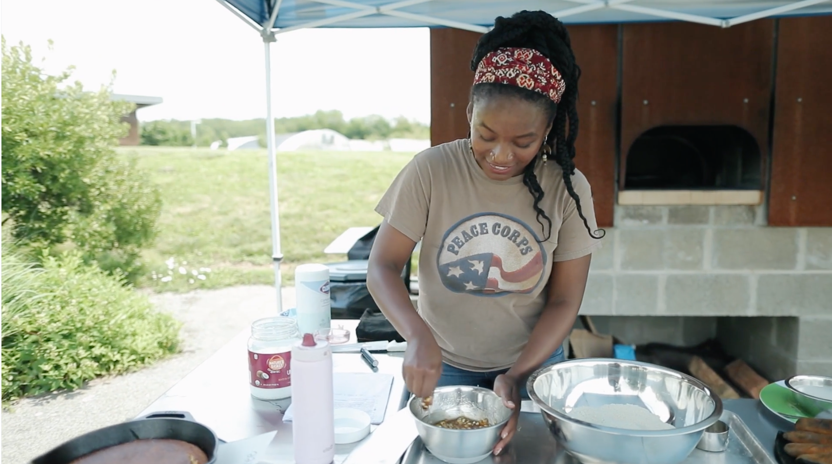 Photo of a Black woman in a Peace Corps shirt, stirring food in a metal bowl, by the Eden Hall Campus Community Bread Oven