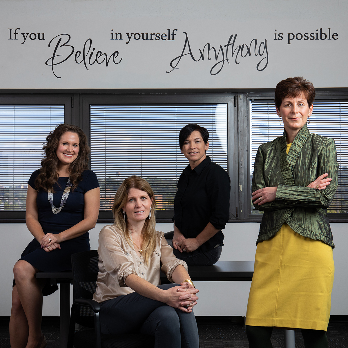 Photo of four women in professional clothing posing in front of a wall upon which is written the quote, "If you believe in yourself anything is possible"