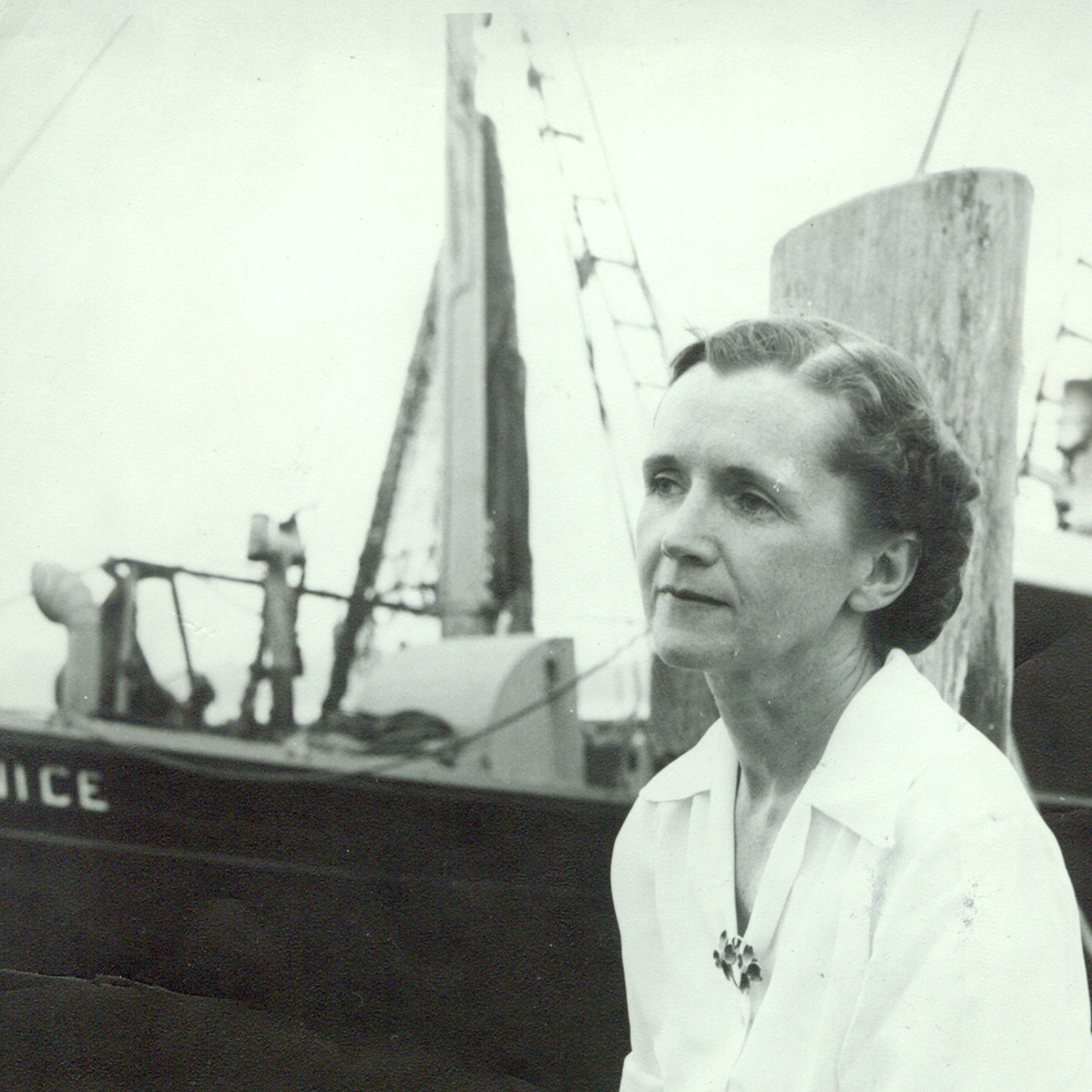Black and white photo of Rachel Carson sitting on a dock, with a pencil and paper in her hand a boat in the background