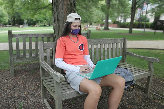 Photo of a Chatham University student in a mask, outside, sitting on a bench and working on her compuer.