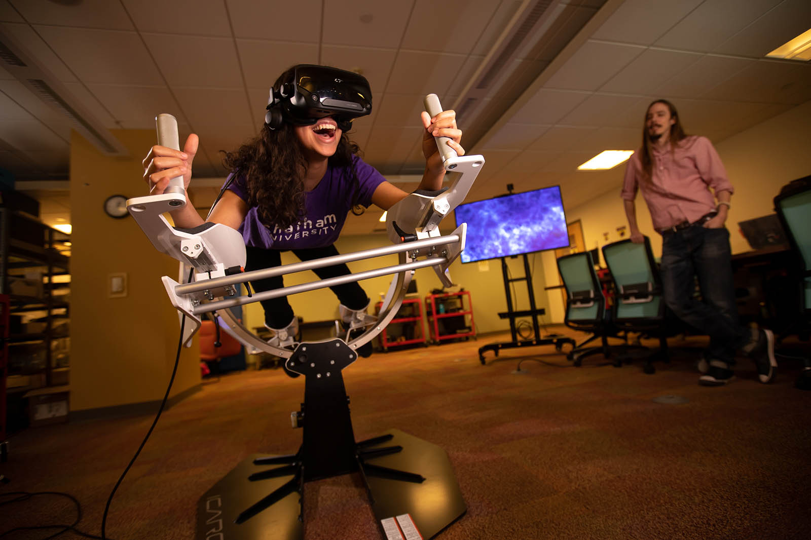 Photo of a Chatham University Immersive Media student in a virtual reality machine, smiling, while a professor sits near.