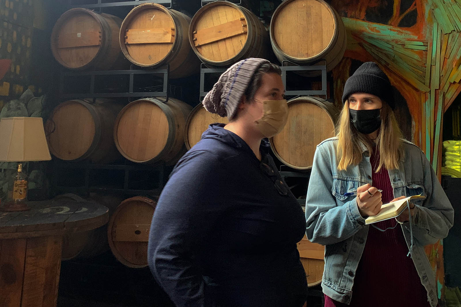 Photo of two people wearing masks, one with a notebook and pen in hand. Behind them there are barrels.