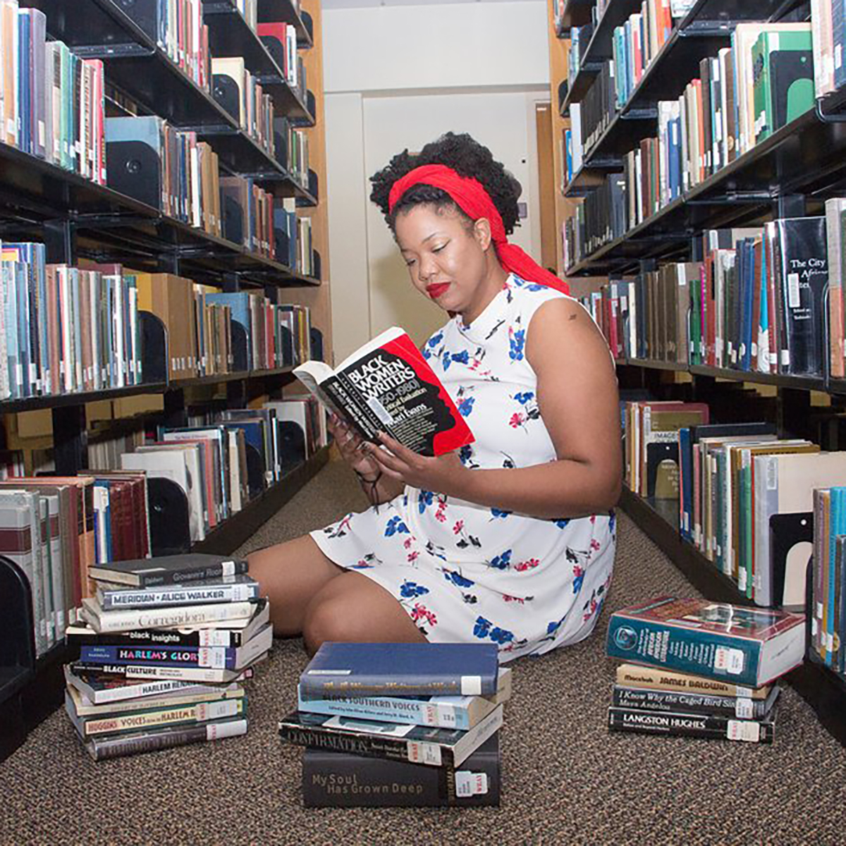 Photo of Caitlyn Hunter, a young Black woman, seated between two library bookshelves with a pile of books in front of her. She is reading a book titled "Black Women Writers, 1950-1980"