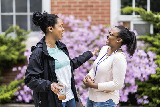 Photo of two women stand together laughing outside of a redbrick Chatham University academic building