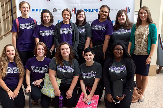 Photo of the Chatham Chamber Choir, wearing purple Chatham University t-shirts, posing together for a photo with the choir director