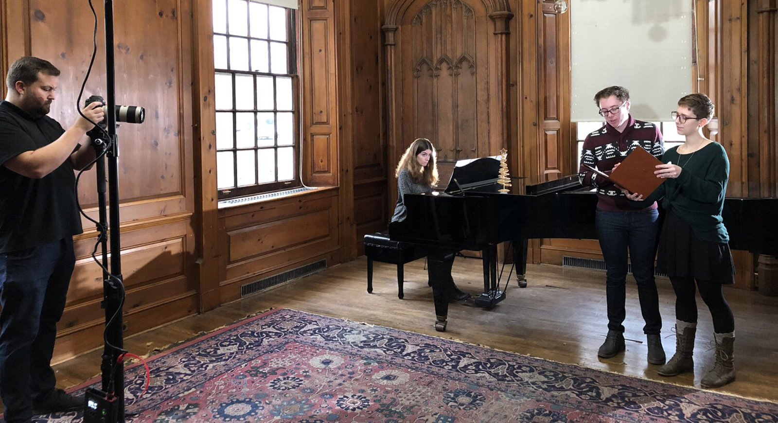 Photo of two students standing and singing, with a woman playing a grand piano behind them. They are being filmed by a person on the left of the photo.
