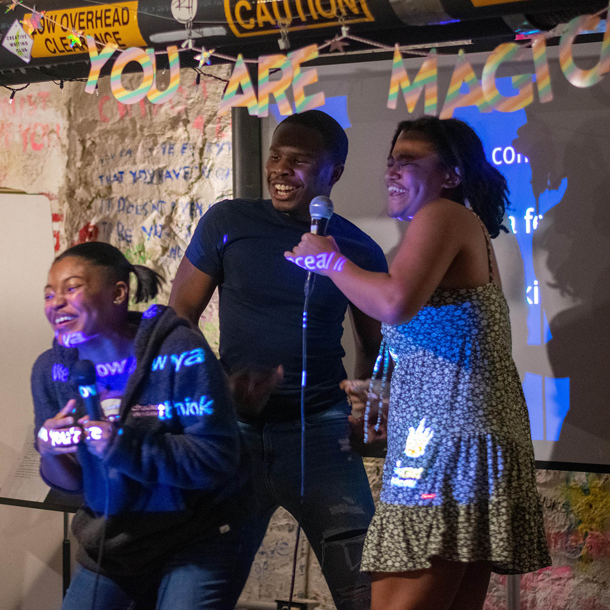 Photo of four Black students laughing and sining on stage with microphones during BIPOC karaoke night. There is a sign behind them that says YOU ARE MAGIC.