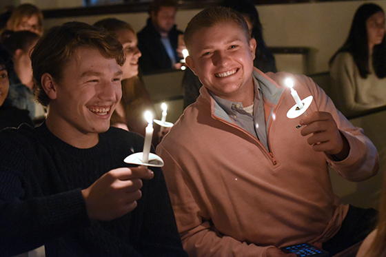 Photo of two Chatham University students smiling while holding candles in a candlelit chapel service on campus. 