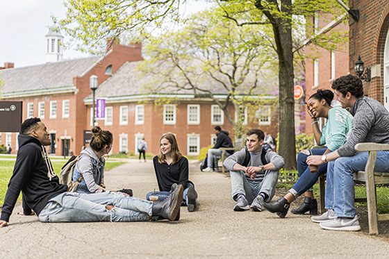 Photo of a group of six Chatham University students sitting outside a red brick academic building on the ground and benches talking